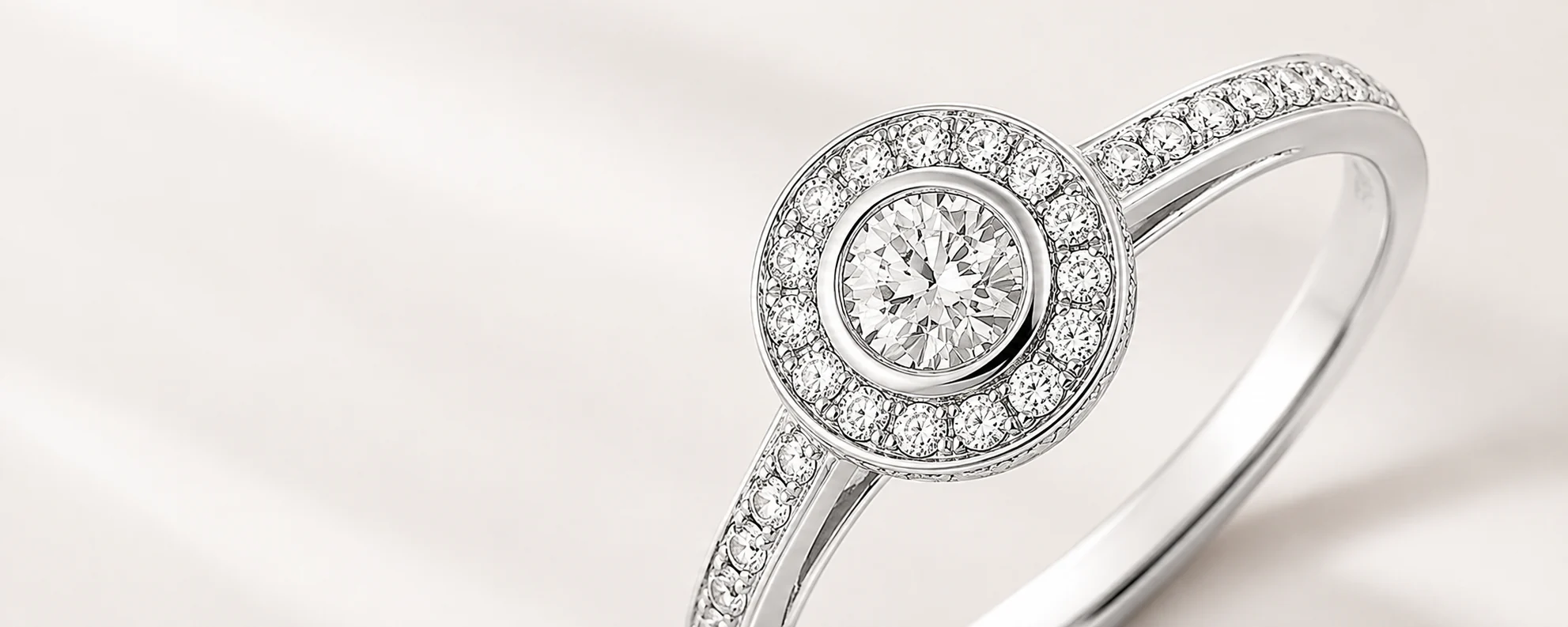 A white gold halo engagement ring — a bezel-set round diamond framed by a diamond halo, with pavé-set shoulders, on cream silk